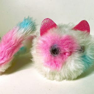 May include: A plush cat-shaped toy made of white, pink, and light blue faux fur. The toy features two bright pink glittery ears, dark circular eyes, and a small pink nose. A long, fluffy, multi-colored tail extends from the side of the round body.