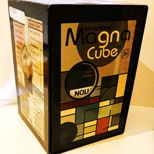 May include: A black box featuring the words "Magna Cube" in large, yellow letters. The box has a geometric design in shades of brown, green, red, and blue. The box is labeled "IQ Collection" and is recommended for ages 8 and up.