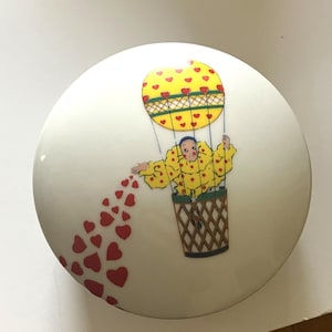 May include: A round, white decorative container with a whimsical design. A yellow hot air balloon with red hearts carries a figure in a yellow polka-dot outfit. Red hearts trail from the figure's hand.