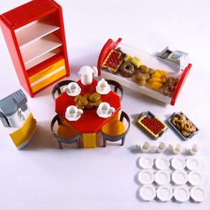 May include: A collection of miniature toy bakery and cafe items. Includes a red cabinet, a red table with chairs, a display case with pastries, and various dishes and cups. The set is designed with bright colors and detailed features.