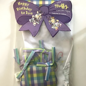 May include: A purple bow with the text "Happy Birthday to You" and "The Muffy Collection Tablecloth & Cushions" is displayed on a white background. A green, blue, and purple plaid cushion with matching ribbon ties is also shown.