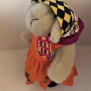 May include: A plush bunny wearing an orange dress and a headscarf with a black and yellow checkered pattern. The bunny has a tag that says "Check Mates". The bunny is standing on a white surface.