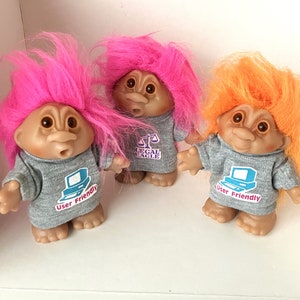 May include: Three troll dolls with pink, orange, and pink hair wearing gray t-shirts with different designs. The pink troll on the left has a computer screen design that says "User Friendly". The pink troll in the middle has a scale design that says "Legal Eagle". The orange troll on the right has a computer screen design that says "User Friendly".