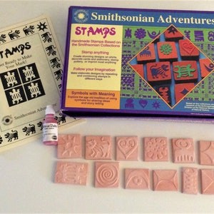 May include: A set of 12 pink rubber stamps with various designs inspired by the Smithsonian Collections. The stamps are arranged in two rows of six. The box for the stamps is purple and features the Smithsonian Adventures logo and the text "STAMPS".