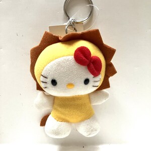 May include: A plush Hello Kitty keychain dressed as a lion. The keychain is yellow with a white face and red bow. The lion mane is brown and the tail is white.