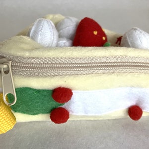 May include: A plush white and green zippered pouch shaped like a strawberry shortcake. The pouch has a yellow strap and a zipper pull.
