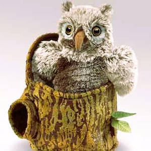 May include: A plush owl toy with large, expressive eyes and a curved beak, perched inside a decorative tree stump. The stump is brown and green with a textured pattern, and a small green leaf is attached to the side.