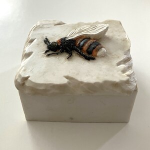 May include: A white ceramic box with a raised image of a bee with black and orange stripes. The bee is in a realistic style and has detailed wings.