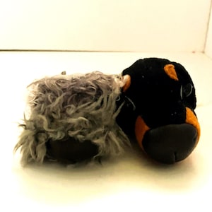 May include: Two plush animal heads positioned side by side. One head is covered in long, shaggy gray fur with a dark nose. The other head features short black fur with tan markings around the eyes and muzzle, and a smooth, dark, rounded nose.