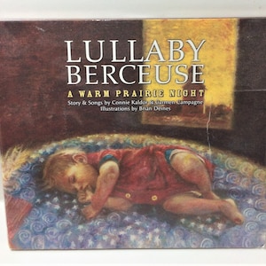 May include: A book titled "Lullaby Berceuse: A Warm Prairie Night" with illustrations of a baby sleeping on a bed with a blue and white patterned blanket. The book features a story and songs by Connie Kaldor and Carmen Campagne, and illustrations by Brian Deines.