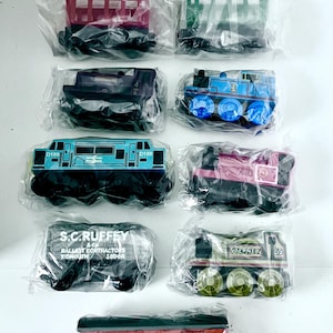 May include: A collection of wooden toy train cars and animal figures, each individually packaged. The train cars are various colors, including blue, red, and green, with printed details and text. The animal figures are brown and black.