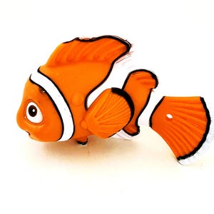 May include: Orange and white clownfish figurine with black accents. The figurine has a large, round body and a small, pointed mouth. It has a large, round eye and a small, black fin on its back.