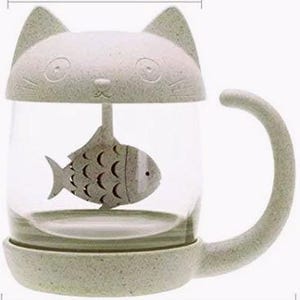 May include: A clear glass mug featuring a light green speckled lid shaped like a cat head with ears. Inside the mug hangs a light green speckled fish-shaped tea infuser. The mug has a matching light green speckled base and a curved cat tail handle.