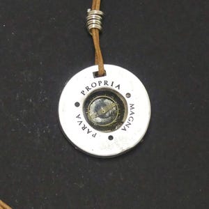Peut inclure: Un collier pendentif argenté avec un design de boussole. Le pendentif porte les mots "PROPRIA PARVA MAGNA". Il est doté d'un cordon en cuir marron et d'un fermoir argenté.