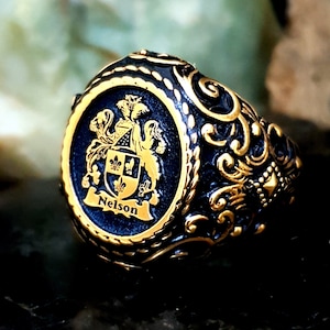 Könnte beinhalten: Ein goldener und schwarzer Ring mit einem detaillierten Design, das ein Wappen mit dem darunter eingravierten Namen "Nelson" zeigt. Der Ring hat einen schwarzen Hintergrund und goldene Akzente.