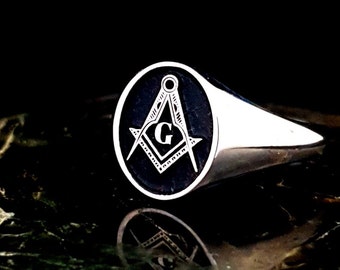 May include: A silver ring with a black oval-shaped setting featuring a silver Masonic symbol with a 'G' in the center.