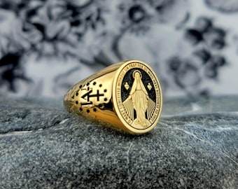 May include: A gold-colored ring with a religious design. The ring features a detailed engraving of a religious figure and text. The ring has a polished finish and is set against a textured background.
