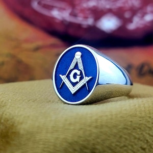 May include: A silver-colored ring with a blue oval face. The face features a white Masonic symbol with the letter "G" in the center. The ring has a polished, reflective surface.