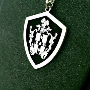 May include: A silver pendant in the shape of a shield with a black enamel background. The shield features a white crest with a cross and four small circles. The pendant is on a silver chain.