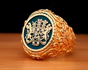 May include: A gold ring with a green enamel crest featuring a coat of arms. The ring has a detailed, ornate design.