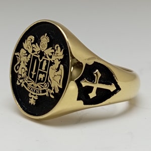 May include: A gold signet ring with a black enamel oval top featuring a coat of arms with the word "WAYNE" underneath. The ring also has a black enamel shield with a gold cross on the side.