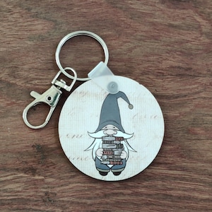 May include: A round keychain featuring a cartoon gnome holding a stack of books. The gnome wears a gray hat and outfit, with a long white beard. The keychain has a silver clasp and ring.
