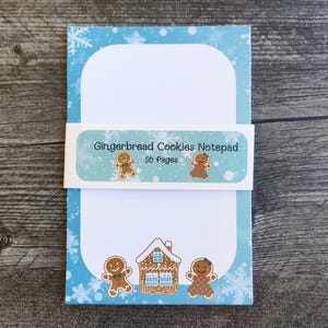 May include: A notepad with a light blue border featuring snowflakes and gingerbread men. The notepad has a white interior with a label that reads "Gingerbread Cookies Notepad" and "50 Pages".