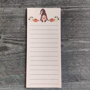 May include: A rectangular notepad with horizontal lines. The top of the notepad features a whimsical illustration of a gnome wearing a brown hat, flanked by orange and white pumpkins. The notepad is on a wooden surface.