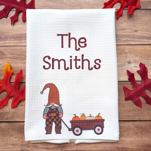 May include: A white waffle-weave kitchen towel featuring the maroon text The Smiths above a graphic of a gnome wearing a plaid hat and overalls, pulling a small wagon filled with four pumpkins in shades of orange, yellow, and white.
