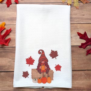 May include: A white waffle-weave kitchen towel featuring a printed graphic of a gnome wearing a striped hat, sitting on a hay bale while holding a pumpkin. The design includes several scattered autumn leaves in shades of red and brown.