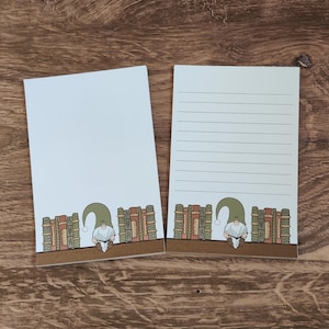 May include: Two white notepads with a brown border. One notepad is blank, the other has horizontal lines. Both have a whimsical illustration of a gnome reading in front of a bookshelf.