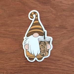 May include: A sticker featuring a gnome with a white beard, holding an iced coffee. The gnome wears a brown and tan striped hat and matching boots. The iced coffee has a straw and a swirl design.