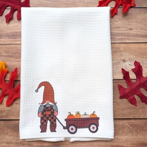 May include: A white waffle-weave kitchen towel featuring a printed graphic of a gnome wearing a brown patterned hat and plaid overalls, pulling a small brown wagon filled with four orange and white pumpkins on a wooden surface.