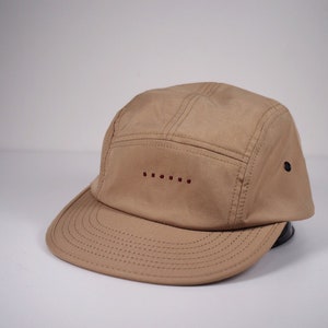 May include: A tan 5-panel cap with a red embroidered design on the front. The cap has a curved brim and a black button on the side.