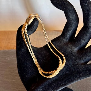 May include: Gold-toned multi-strand bracelet with a rectangular clasp. The bracelet features three delicate chains and a textured center chain. The jewelry is displayed on a black hand-shaped display.