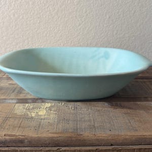 Può includere: Una ciotola in ceramica azzurra di forma rettangolare arrotondata, con piccole maniglie. Poggia su una superficie di legno. Il colore della ciotola è un blu-verde tenue e smorzato.