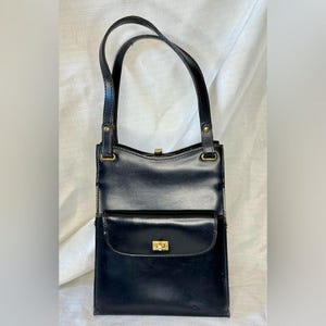 Puede incluir: Bolso de mano de cuero negro con forma rectangular y bolsillo delantero. El bolso tiene dos asas y un cierre dorado. El bolso mide aproximadamente 25 cm de alto y 20 cm de ancho.