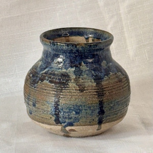 Pode incluir: Um pequeno vaso de cerâmica redondo com uma boca larga e um pescoço estreito. O exterior apresenta um padrão texturizado de nervuras horizontais com um esmalte que transita de um azul profundo e brilhante para uma base castanha e bege.