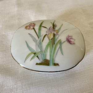 May include: An oval-shaped white porcelain trinket box featuring a delicate hand-painted floral design of purple and pink irises with green stems and leaves. The lid is finished with a thin gold-toned metallic trim along the outer edge.