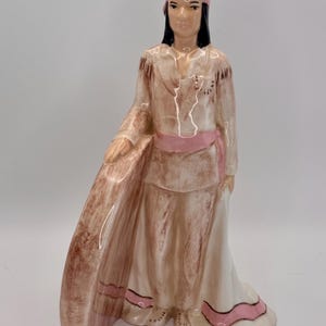 Puede incluir: Figura de cerámica de una mujer nativa americana con vestimenta tradicional. La figura lleva un vestido claro con detalles rosas, una diadema y cabello negro. La figura tiene un acabado detallado y pintado a mano.