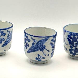 May include: Three small, white ceramic cups with blue floral and crane designs. The cups have a rounded shape with a slightly flared rim and a small footed base. The blue designs are hand-painted, giving each cup a unique look.