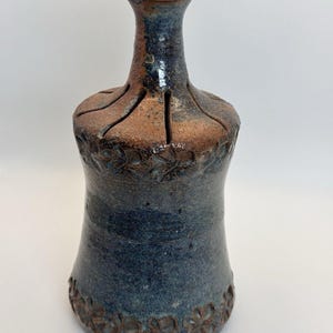 May include: A ceramic vase with a narrow neck and a flared base, featuring a mottled blue and brown glaze. The upper shoulder and lower body are decorated with embossed floral patterns, and the neck has four vertical slits cut into the top.