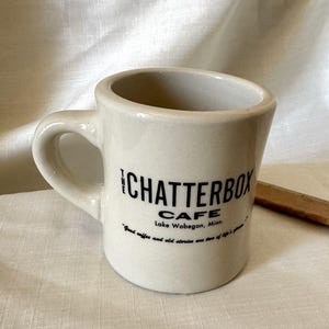 Op de afbeelding: Een crèmekleurige keramische mok met een dik handvat en zwarte tekst die luidt THE CHATTERBOX CAFE Lake Wobegon, Minn. en Good coffee and old stories are two of life's pleasures. De mok heeft een gladde, glanzende afwerking en een brede opening.