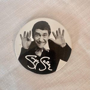 May include: A round, black and white button featuring a smiling man with his hands up. The man is wearing a suit and tie. The button has the name "Spy See" in a white, cursive font.