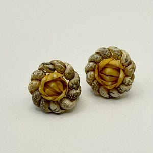 Könnte beinhalten: Ein Paar handgefertigte Ohrringe mit floralem Design. Jeder Ohrring hat eine zentrale, hellgelbe Blume, die von einem Ring aus kleinen, beige-braunen Muscheln umgeben ist. Die Ohrringe sind vor einem weißen Hintergrund platziert.