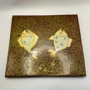May include: Square ceramic tile with a speckled brown and green background. Two stylized fish in cream with blue and orange accents are connected by a thin, curved line. The fish have star-shaped details and are facing each other.