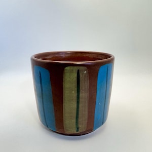May include: A cylindrical ceramic planter with a smooth brown base finish. The exterior features vertical painted stripes in alternating bright blue and muted olive green, each accented with a thin, dark vertical line running through the center.