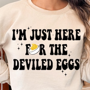 Puede incluir: Una sudadera beige con el texto "I'm Just Here For The Deviled Eggs" en negro. Se incluye una pequeña ilustración de un huevo relleno en el texto.