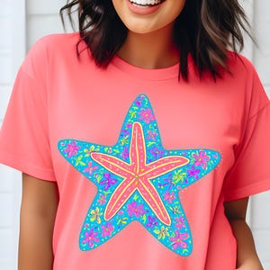 May include: A coral-colored t-shirt featuring a large starfish design. The starfish has a pink center with yellow accents and is outlined in blue. The starfish is decorated with colorful flowers, creating a tropical theme.