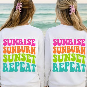 May include: Two white sweatshirts with the words "SUNRISE SUNBURN SUNSET REPEAT" in bright pink, orange, green, and teal lettering. The sweatshirts are worn by two people with blonde hair, each wearing a pink hair clip. The background is a beach scene.